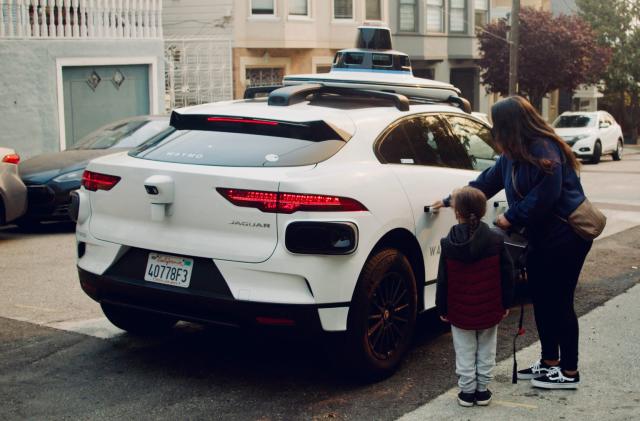 A woman and a child and are standing next to a white autonomous vehicle, in the process of pulling a door open. 