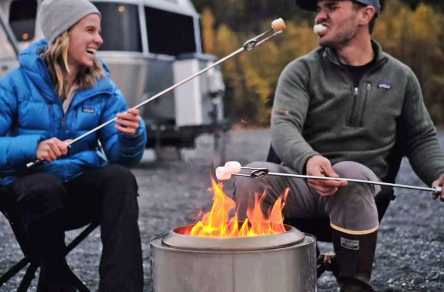 A Solo Stove firepit burns in an outdoor setting as two people toast marshmallows in its flames. A camper trailer is in the background.  