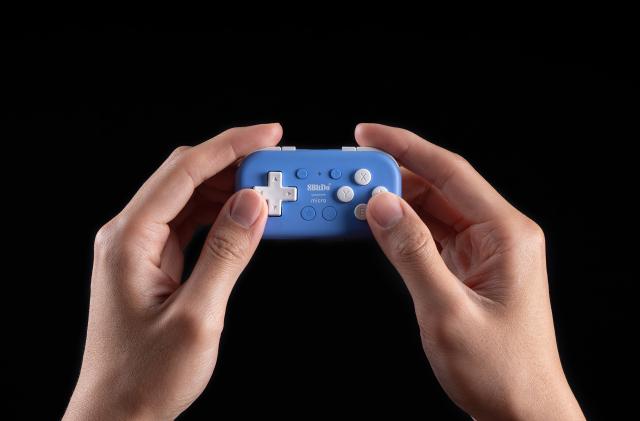A pair of hands holding a tiny blue 8BitDo Micro gamepad in front of a black background.