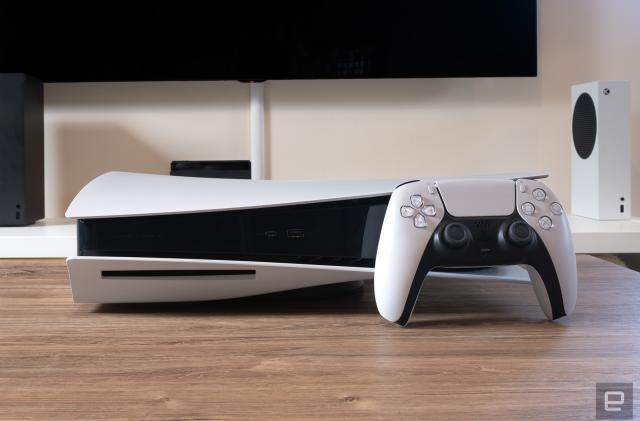 The white and black PlayStation 5 console is on a wooden coffee table with a TV and Xbox in the background. A PlayStation controller sits in front of the console. 