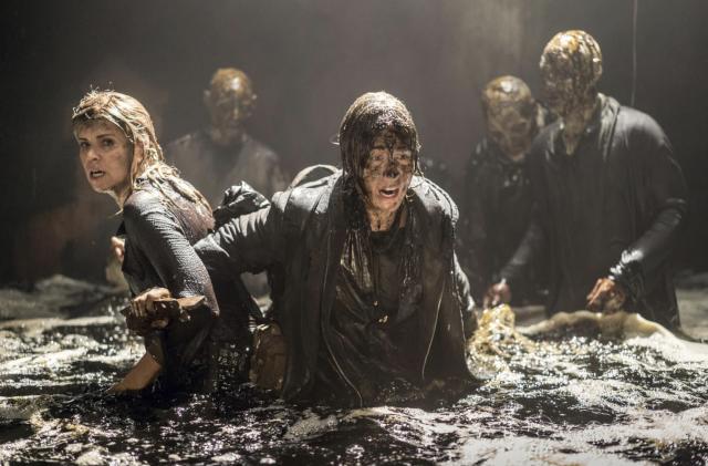 Two women immersed in waist-high muddy water back to back, being chased by zombies with rotting faces.