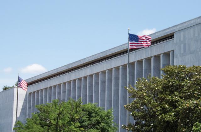 Outside view of a regal Washington, DC government building with a tall American flag and tree tops in front.