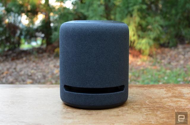 Amazon's Echo Studio Speaker on a wooden table outdoors in front of a bank of trees. 