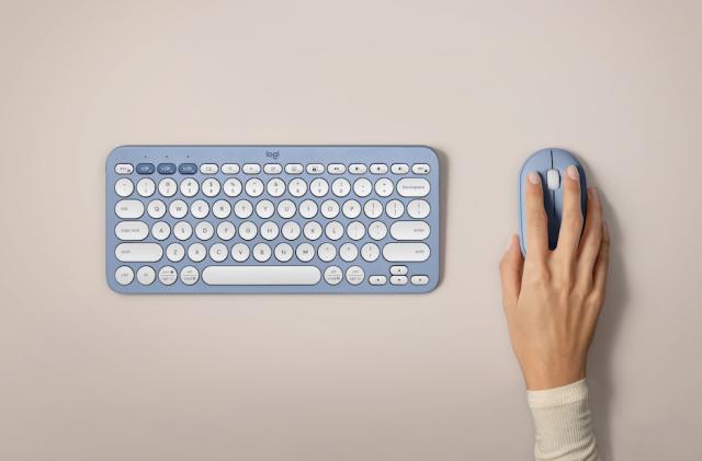 A blue Logitech Pebble 2 keyboard and mouse seen from above on a blank table top with a hand grasping the mouse.