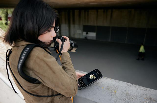 A photographer looks away from their camera toward their phone, which displays a phot-editing app.