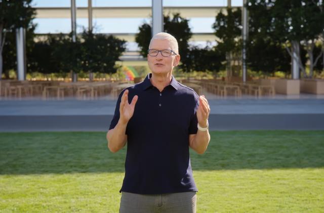 Screenshot of Tim Cook presenting at Apple's September 2022 iPhone launch event. He stands outdoors at Apple Park, on green grass in front of the "spaceship" building, holding two hands in front of him to emphasize a point.