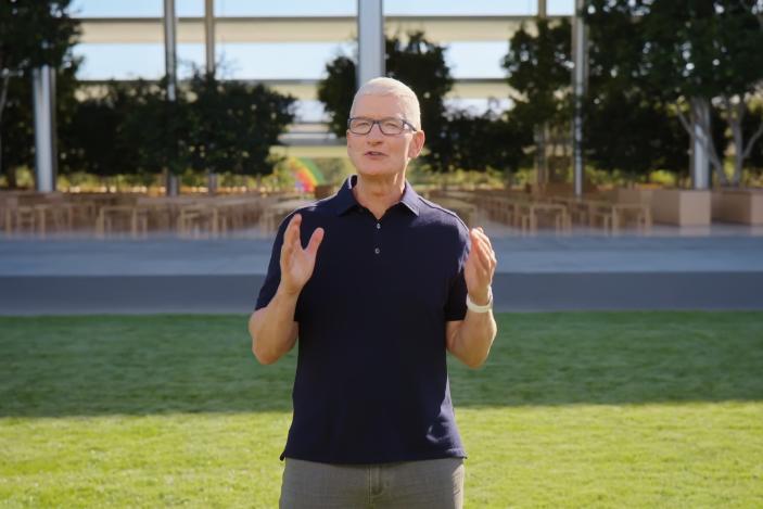 Screenshot of Tim Cook presenting at Apple's September 2022 iPhone launch event. He stands outdoors at Apple Park, on green grass in front of the "spaceship" building, holding two hands in front of him to emphasize a point.