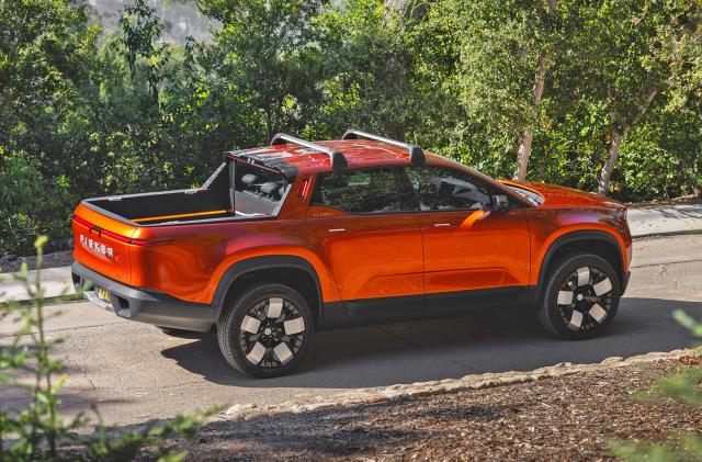 A Fisker Alaska 2023 pickup in burnt orange color is seen driving along a wooded road on a hillside, with trees flanking the road.